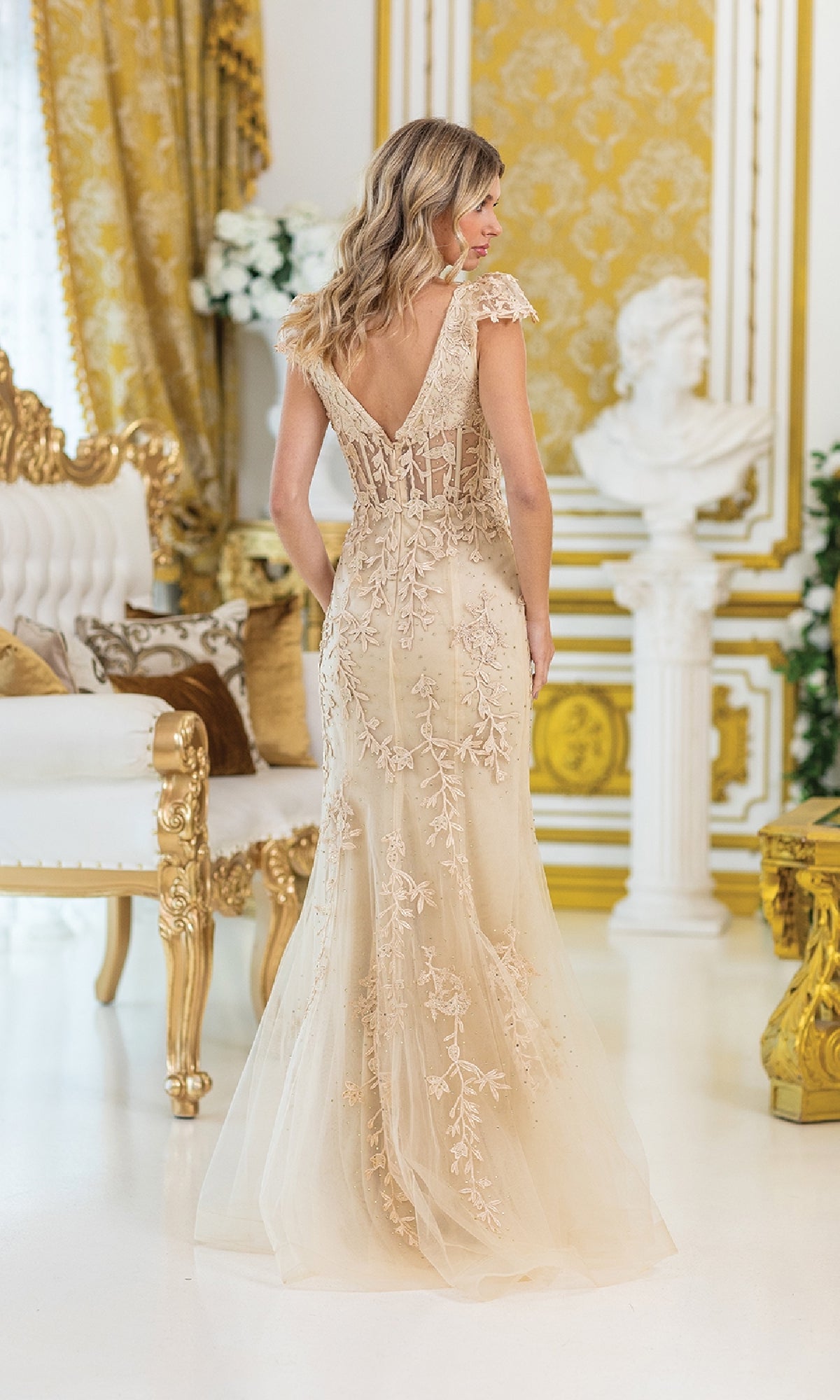 A woman wearing the Cap-Sleeve Long Prom Dress: Dancing Queen 4447, a floor-length beige gown with floral embroidery, stands in an ornate room featuring gold and white decor.